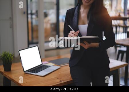 Felice donna d'affari che prende nota con il computer portatile sulla scrivania in legno in ufficio Foto Stock