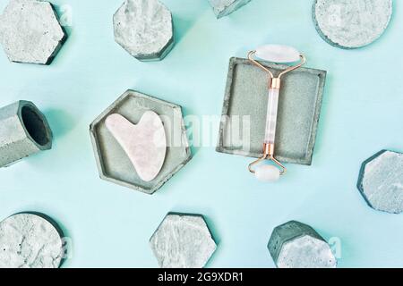 Massaggiatore e rullo di Gua Sha in quarzo rosa su podi in cemento su sfondo blu. Per la cura del viso e del corpo. Disposizione piatta, vista dall'alto. Foto Stock