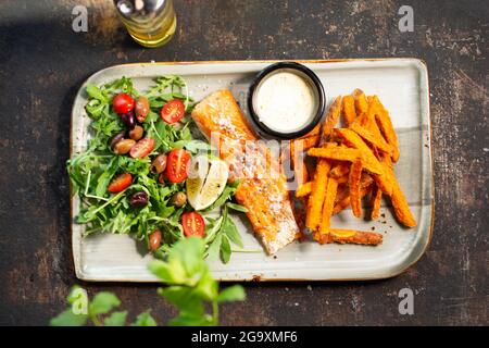 Salmone alla griglia con patatine fritte dolci e insalata. Il cuoco cucina e serve un piatto appetitoso. Il piatto finito servito su un piatto. Proposta di servizio Foto Stock