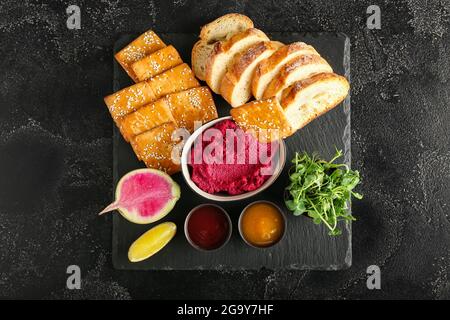 Ciotola con gustoso hummus di barbabietola, pane, cracker, germogli freschi e salse su fondo scuro Foto Stock