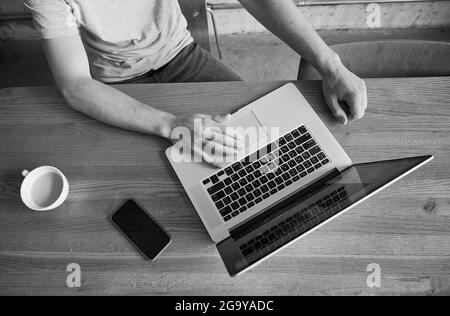 Vista dall'alto delle mani dei maschi che lavorano sul computer portatile in bianco e nero. Aspetto dell'uomo seduto al tavolo con computer portatile, smartphone e tazza di caffè. Concetto di lavoro online. Immagine monocromatica Foto Stock