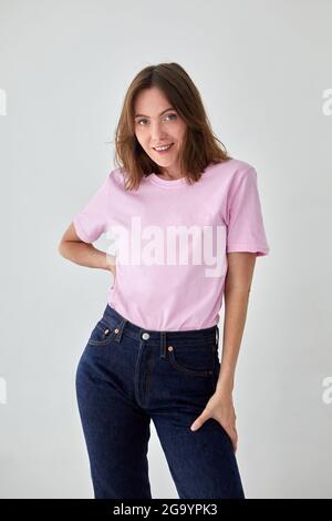 Modello femminile positivo con t-shirt rosa e jeans in piedi con la mano sulla vita su sfondo bianco e guardando la fotocamera Foto Stock