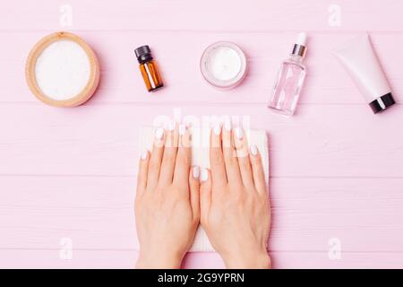 Mani donna con manicure bianco accanto a prodotti cosmetici per la cura delle unghie, scrub al sale marino, olio di cuticola, crema e lozione su sfondo rosa, vista dall'alto. Foto Stock