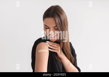 Paziente caucasico di sesso femminile che mostra spalla con aiuto di fascia dopo iniezione di vaccino anti covid su sfondo chiaro Foto Stock