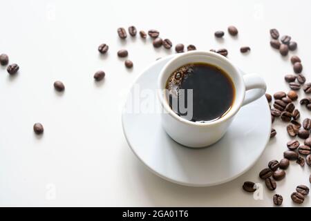 Caffè caldo, nero e forte in una tazza bianca su un tavolo luminoso con alcuni chicchi, spazio di copia, messa a fuoco selezionata, profondità di campo ridotta Foto Stock