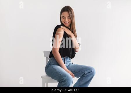 Donna caucasica sorridente che mostra la spalla con l'aiuto della fascia dopo l'iniezione del vaccino del coronavirus su sfondo di studio chiaro Foto Stock