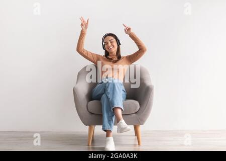 Giovane donna cool seduta in poltrona, ascoltando musica in cuffie wireless, ballando alla sua canzone preferita Foto Stock