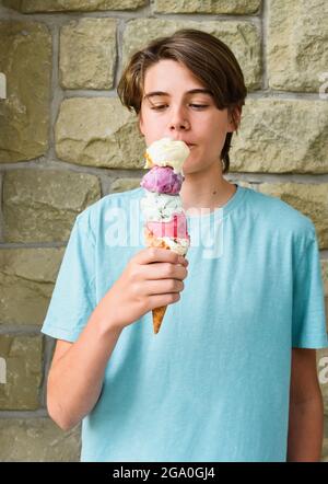 Teen boy che mangia un grande cono di gelato a paletta multipla all'esterno. Foto Stock