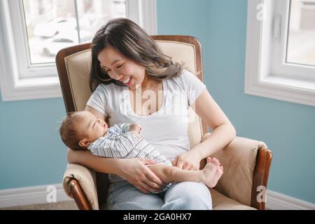 Festa della mamma. Orgogliosa madre cinese asiatica sorridente che tiene dondolo neonato figlio bambino. Stile di vita domestico momento naturale autentico. Incollaggio di m Foto Stock