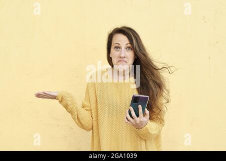 Giovane donna che tiene un telefono cellulare isolato su sfondo giallo avvolge le spalle e gli occhi aperti confusi. Foto Stock