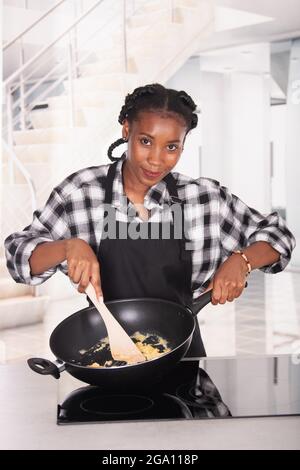 Sorridente afroamericana tenendo una padella profonda e mescolando le uova Foto Stock