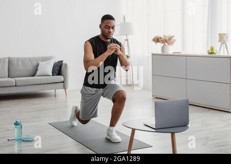 African Man Training al laptop facendo Forward Lunge a casa Foto Stock