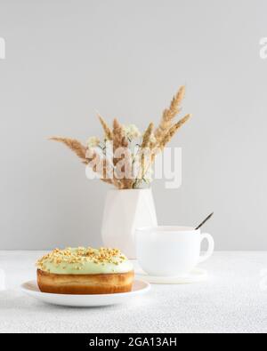 Ciambella al pistacchio e tazza di caffè su un tavolo con fiori secchi in piccolo vaso minimalista sullo sfondo Foto Stock