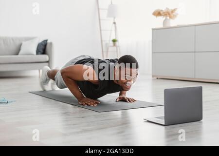 Atletico Guy Africano che si esercita a laptop facendo Push-UPS a casa Foto Stock