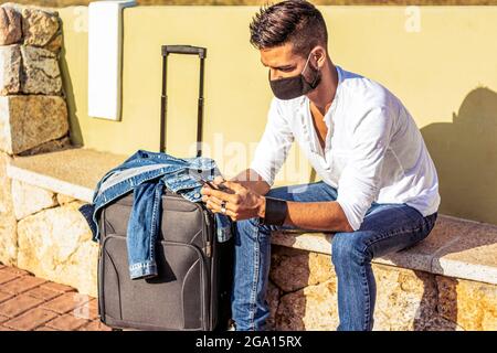 Solo viaggiatore in attesa di trasporto pubblico seduto sulla panca indossando una maschera protettiva nera sicura utilizzando smartphone per passare noioso attesa. Elegante Foto Stock