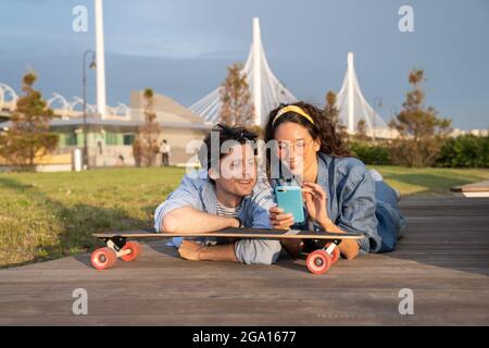 Giovane ragazza elegante mostra il messaggio di testo ragazzo su smartphone sdraiato a bordo lungo in un parco urbano all'aperto Foto Stock