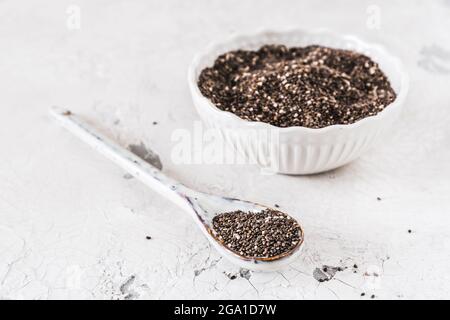 I semi di Chia su un cucchiaio di porcellain bianco davanti ad una ciotola bianca con i semi di chia, mettere a fuoco sul cucchiaio, sfondo bianco, spazio di copia Foto Stock