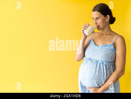 ritratto cute bella gravidanza giovane bella donna graziosa in abito blu floreale tiene un bicchiere da bere di latte strofina tummy su sfondo giallo. Maternità Foto Stock
