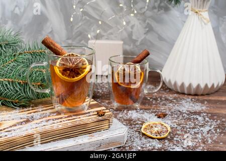 Carta anno nuovo. Due tazze di tè di vetro con bastoncini di limone e cannella sullo sfondo delle ghirlande di Natale, un regalo e rami di un albero di Natale Foto Stock