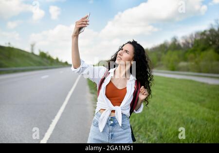 Turbata giovane donna che cammina lungo la strada, alzando la mano con smartphone, alla ricerca di segnale, senza connessione, all'aperto Foto Stock