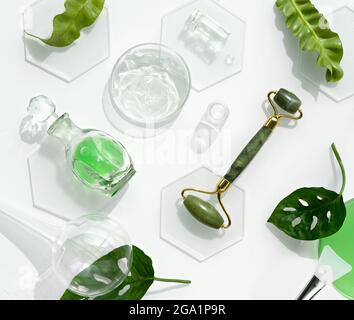 Moisturizer e rullo per la faccia in giada verde con pezzi di ghiaccio. Foglie esotiche di monstera e gocce d'acqua su sfondo bianco. Massaggio viso Foto Stock