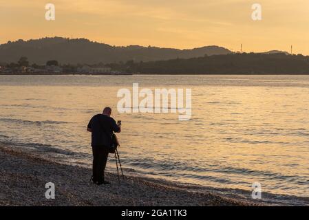 Corfù, Grecia - 7 maggio 2016: Un fotografo con un treppiede che scatta foto del mare e della spiaggia di Acharavi durante il tramonto. Isola di Corfù, Grecia Foto Stock