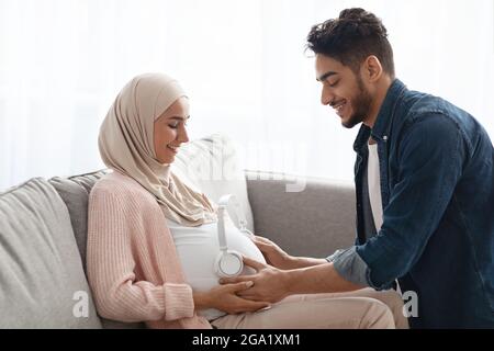 Adorabile marito arabo che mette le cuffie wireless sul venticellano della moglie incinta Foto Stock