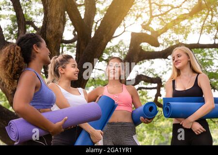 felici giovani donne multietniche amici gruppo di amici rilassarsi e parlare dopo l'esercizio di yoga nel parco al fine settimana mattina. vita dopo covid. Foto Stock