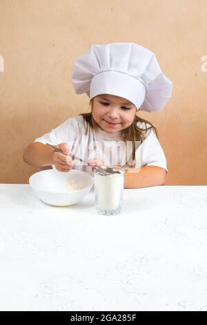 Una bambina vestita come cuoco impasta l'impasto. Cucina bambino stile di vita concetto. Il capretto ama, ha divertimento, studia e gioca nella cucina Foto Stock