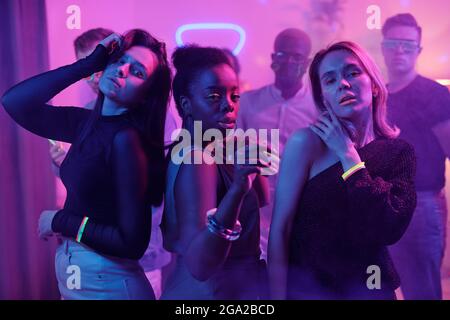 Giovane eccitato uomo in casualwear e discoteca occhiali e suo amici interculturali con bastoni incandescenti che ballano insieme mentre si godono a casa festa Foto Stock