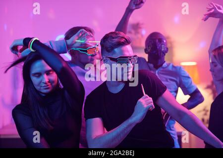 Giovane eccitato uomo in casualwear e discoteca occhiali e suo amici interculturali con bastoni incandescenti che ballano insieme mentre si godono a casa festa Foto Stock