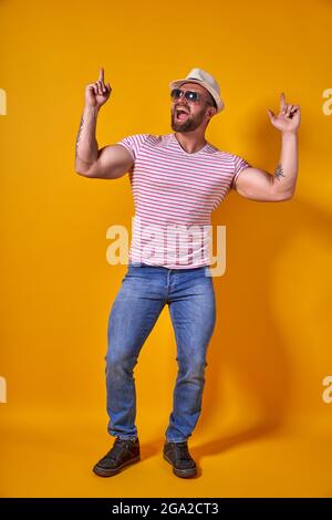 Ritratto di ottimista funky ragazzo danza indossare estate look cappello di paglia, occhiali da sole isolato su sfondo giallo Foto Stock