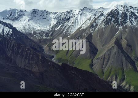 Antenna di pendii di antica roccia sedimentaria e vulcanica con vegetazione verde e cime innevate all'orizzonte, situata nei Tradi... Foto Stock