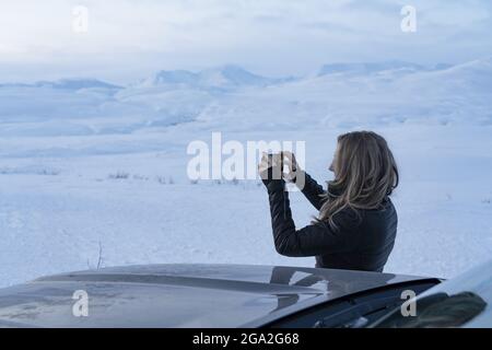 Donna viaggiatore in piedi accanto ad un'auto che prende una fotografia con il suo cellulare del paesaggio nevoso lungo la Haines Highway; Yukon, Canada Foto Stock