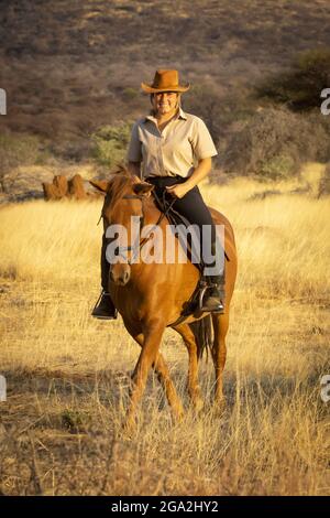 Donna a cavallo (Equus ferus caballus) attraverso il cespuglio sulla savana con la calda luce del tramonto, guardando la macchina fotografica e sorridendo il G... Foto Stock