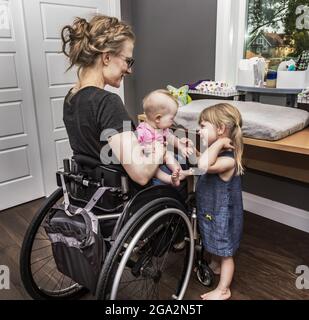 Una madre in sedia a rotelle cambia il pannolino del bambino mentre la sorella grande aiuta divertendo il bambino; Edmonton, Alberta, Canada Foto Stock