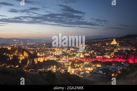 Crepuscolo cade sulla capitale di Tbilisi con le sue luci della città che mettono in risalto la Cattedrale della Santissima Trinità di Tbilisi sulla destra, il Ponte di... Foto Stock