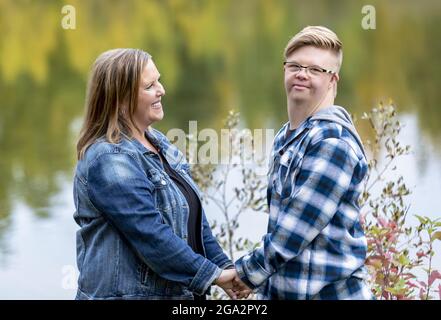 Un giovane uomo con la sindrome di Down che tiene le mani con la madre e guardando la macchina fotografica mentre si godono l'azienda dell'altro in un parco della città... Foto Stock