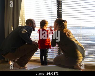 Una giovane madre e un padre si accovacciano dalla loro figlia toddler per guardare insieme una finestra; White Rock, British Columbia, Canada Foto Stock