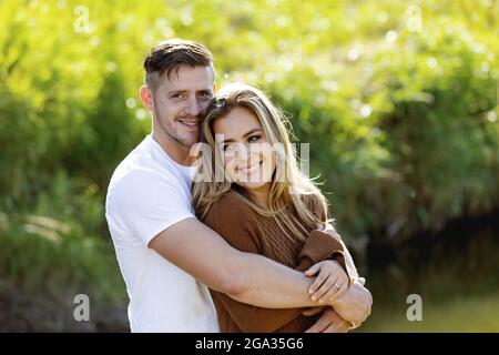 Scatto corto di una giovane coppia sposata sorridente allegro e abbracciandosi mentre si levano in piedi insieme in un parco della città durante il giorno Foto Stock