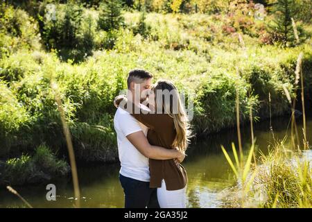 Marito e moglie trascorrono del tempo di qualità insieme e baciano all'aperto vicino a un ruscello in un parco cittadino; Edmonton, Alberta, Canada Foto Stock