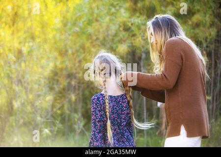 Una madre e una figlia che trascorrono tempo di qualità insieme all'aperto e la mamma sta brasando i suoi lunghi capelli in un parco cittadino durante la stagione autunnale Foto Stock