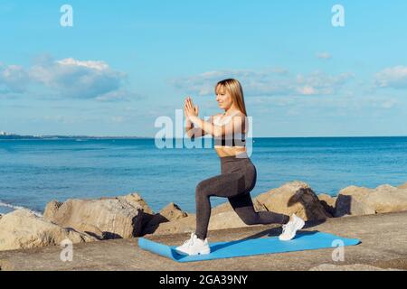 Giovane donna fitness in abbigliamento sportivo facendo affondo esercizi durante l'allenamento all'aperto sulla spiaggia, molo sul mare. Stile di vita attivo e assistenza sanitaria. Copia Foto Stock