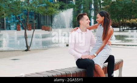 Romantica coppia multirazziale che ha una data: Donna sussurrando qualcosa nell'orecchio del ragazzo. Si siedono su una panchina, abbracciano e baciano. Foto Stock