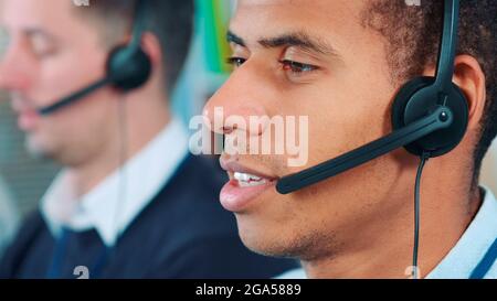 Primo piano di un uomo di razza mista in cuffia che parla con qualcuno in ufficio. Sullo sfondo c'è il suo collega che lavora. Foto Stock