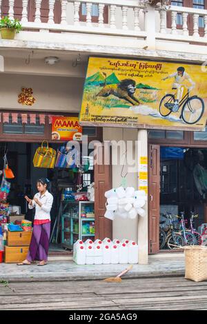 La giovane donna birmana indossava i lunghi si trovava all'esterno del negozio di ferramenta e biciclette con la pubblicità colorata di leone inseguitore uomo in bicicletta, Hsipaw, Myanmar Foto Stock