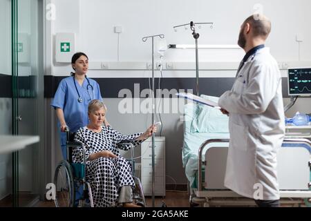 Donna anziana malata ricoverata seduta su sedia a rotelle con sacca fleboclisi iv attaccata al braccio, aiutata dall'infermiere a muoversi. Medico con stetoscopio che controlla il paziente prima dell'intervento chirurgico e che parla di recupero del trattamento. Foto Stock