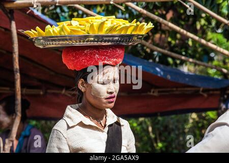 Giovane falciatrice birmana con thanaka sulla faccia che bilancia grande vassoio di frutta di mango sulla testa, Kalaw, Myanmar Foto Stock