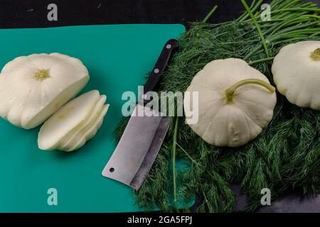 Squash di Pattyan grezzi, un hatchet della cucina, un mazzo di davanzali freschi e un bordo di plastica verde su sfondo nero. Padella con gocce d'acqua Foto Stock