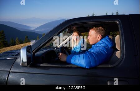 Donna che guarda il marito e sorride mentre l'uomo tiene le mani sul volante e guida l'auto. Giovane uomo e donna che viaggiano insieme Foto Stock